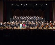 Toda a orquestra, com seus instrumentos, o coro, os solistas, os músicos convidados e o maestro estão posicionados durante o concerto no placo do Teatro Guaíra, com roupas de gala. 