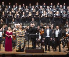 Toda a orquestra, com seus instrumentos, o coro, os solistas, os músicos convidados e o maestro estão posicionados durante o concerto no placo do Teatro Guaíra, com roupas de gala. 