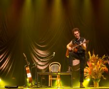 Nando Reis tocando violão no palco. Ao fundo, o cenário conta com um mesa, cadeira, vaso com plantas e outro violão em apoio