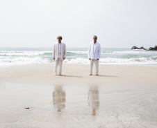 Arnaldo Antunes e Vitor Araújo com roupas claras na beira do mar 