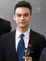 Foto de um músico da cintura para cima vestindo terno e gravata azul. Ele olha para a câmera. 