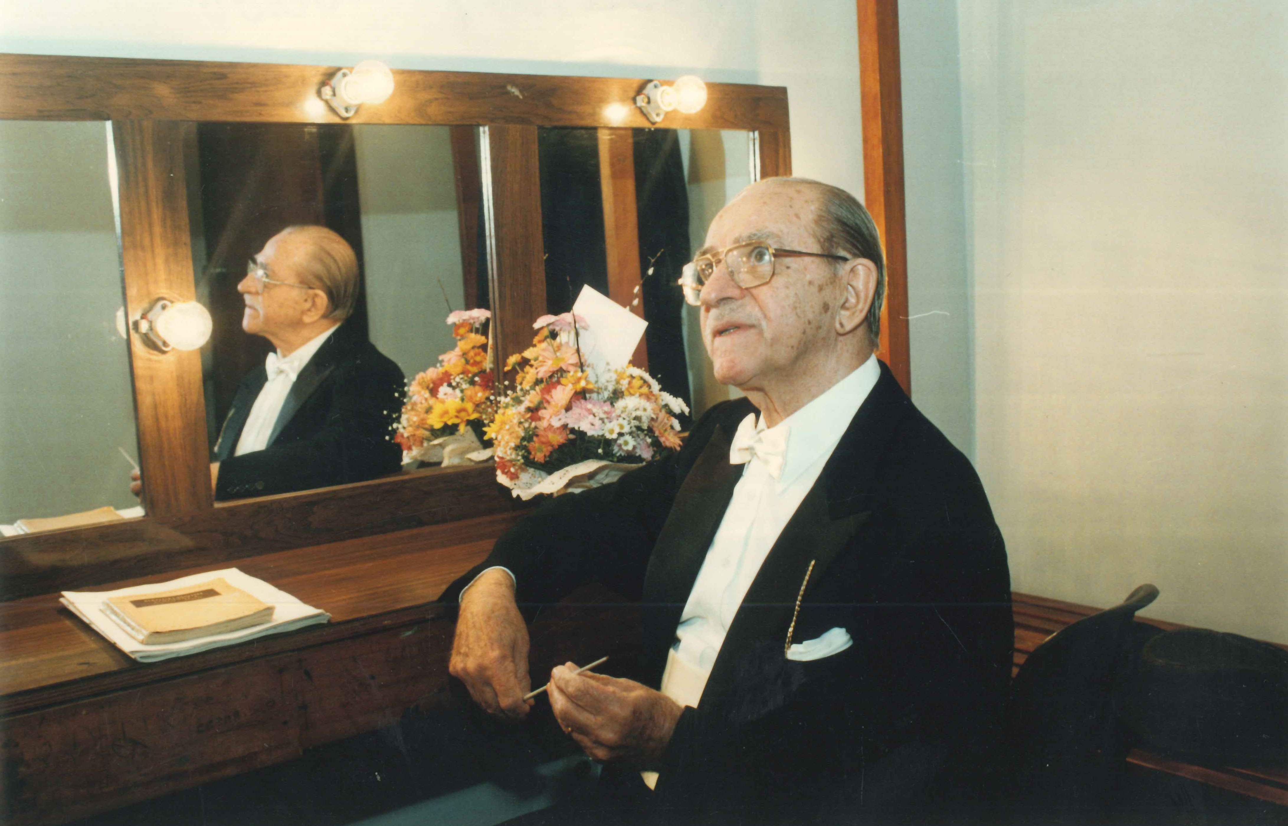 Foto do maestro sentado em um dos camarins. Ele está arrumado para o concerto e segura a batuta entre as mãos