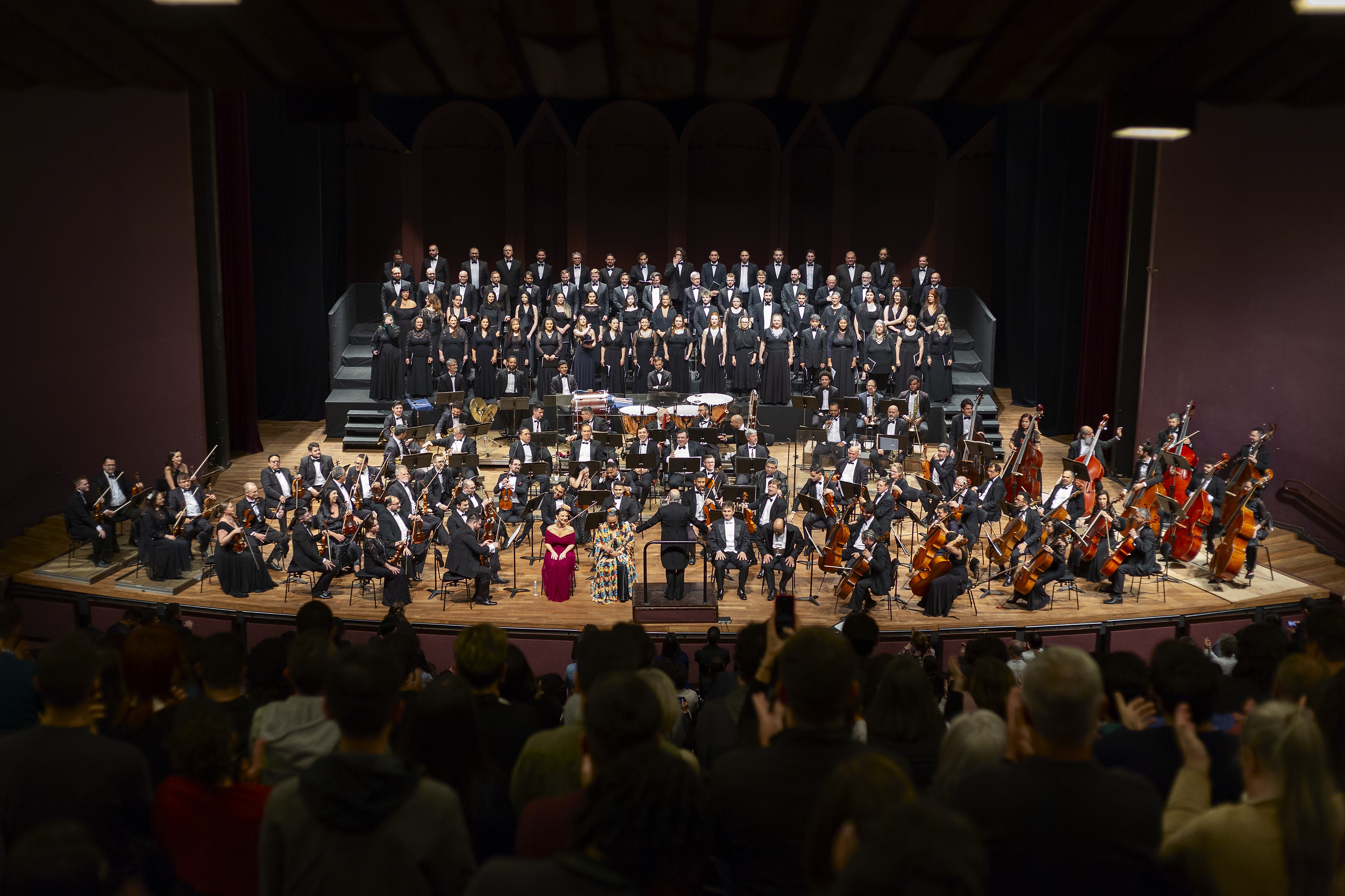 Toda a orquestra, com seus instrumentos, o coro, os solistas, os músicos convidados e o maestro estão posicionados durante o concerto no placo do Teatro Guaíra, com roupas de gala.