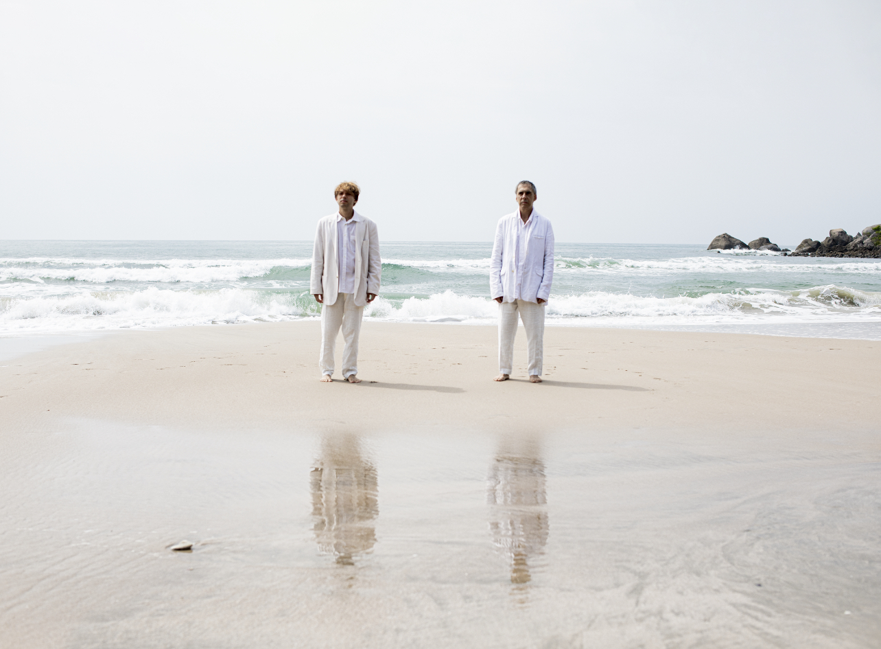 Arnaldo Antunes e Vitor Araújo com roupas claras na beira do mar