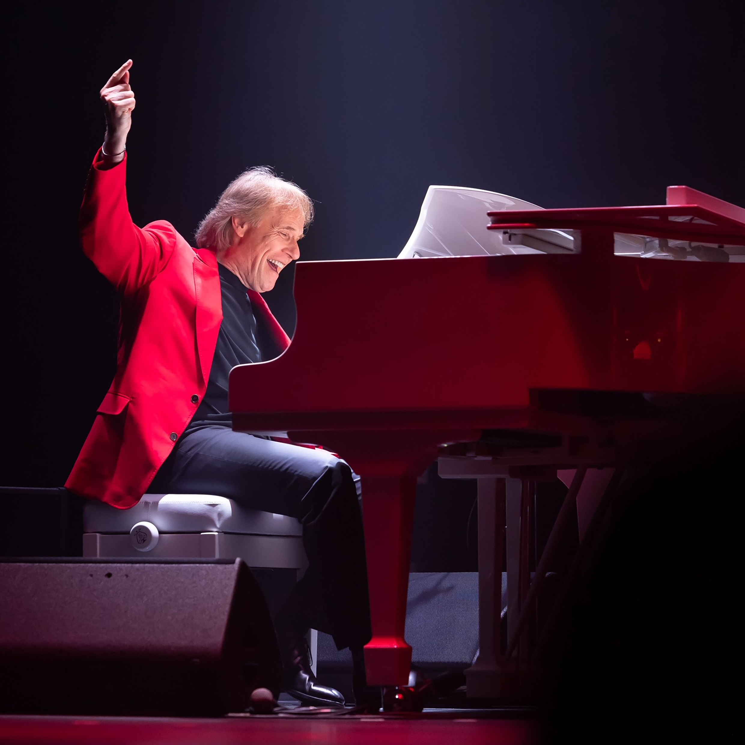O pianista no palco em posição de tocar o piano, com o braço direito erguido em pose alegre.
