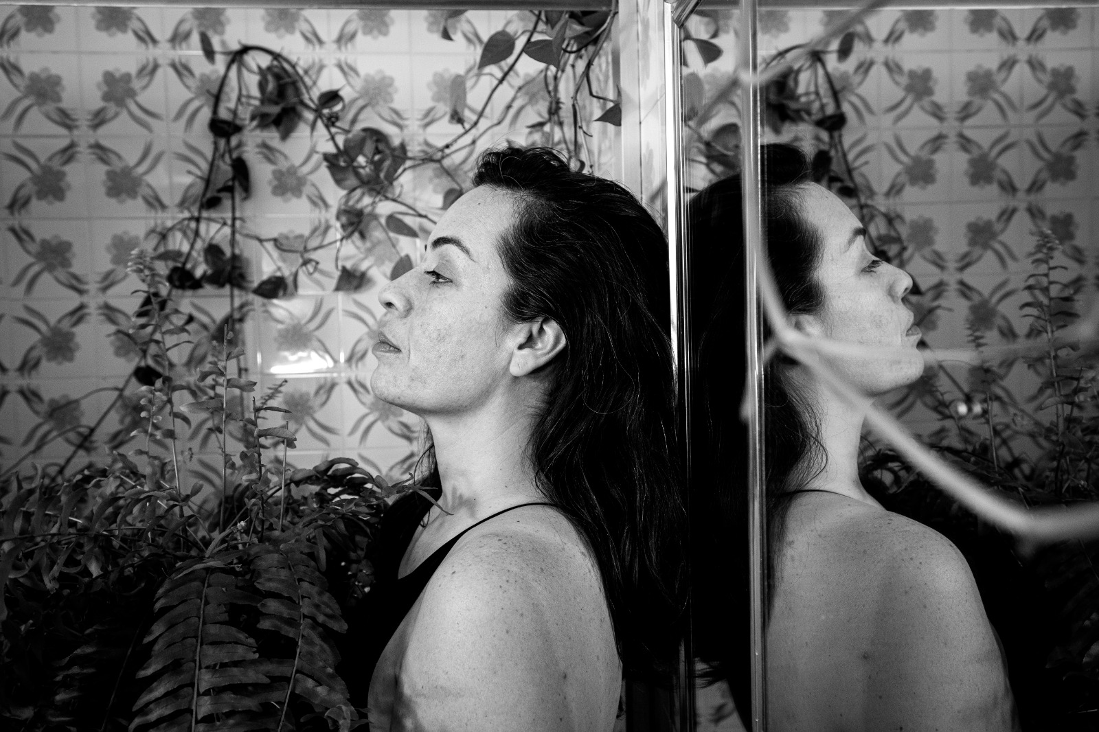 foto em preto e branco, mulher encostada em uma parede com um espelho refletindo sua imagem ao lado. ao fundo, plantas na parede decoram o cenário
