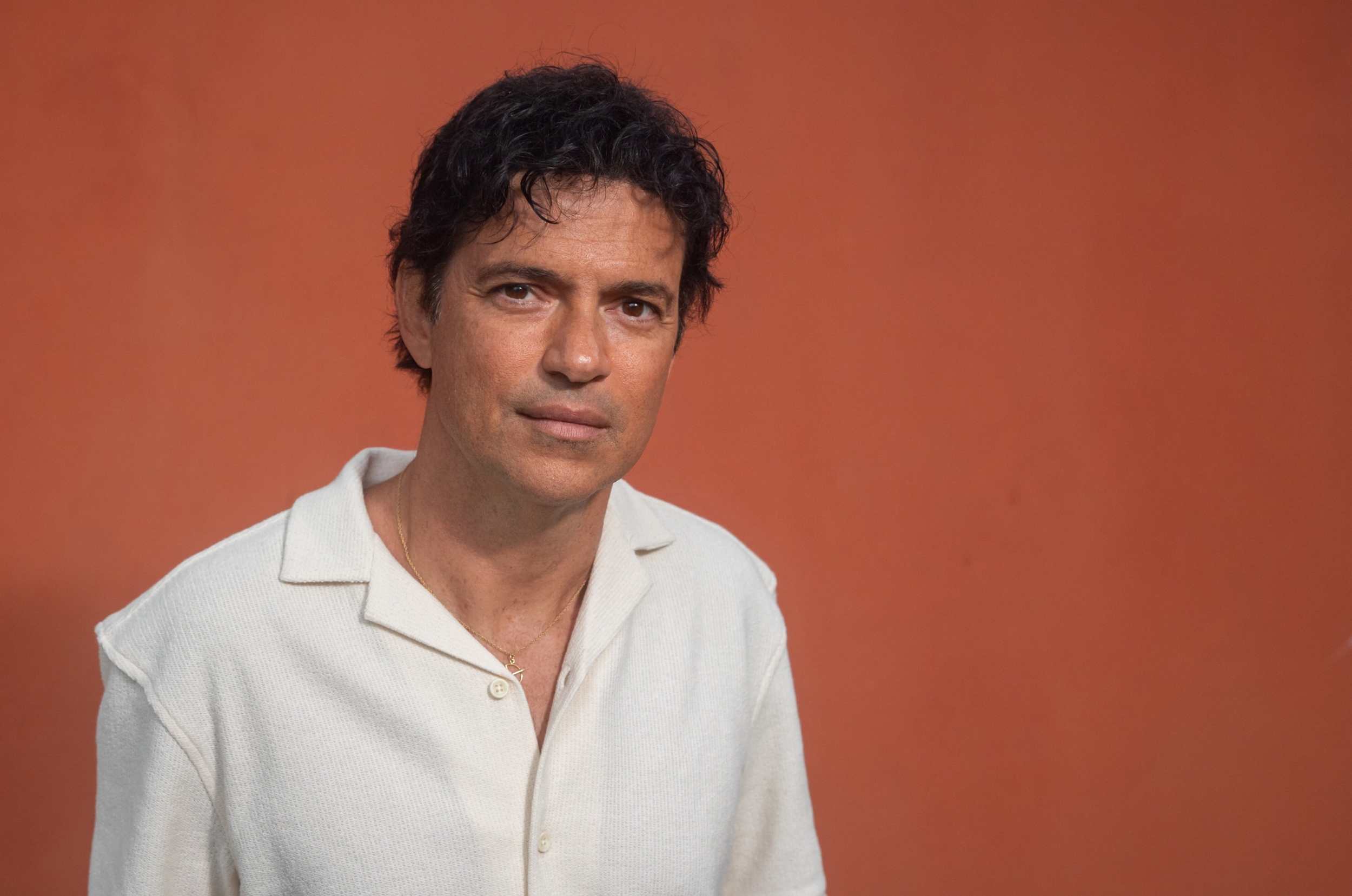 Jorge Vercillo posando para foto em frente a fundo laranja. O cantor veste camisa branca