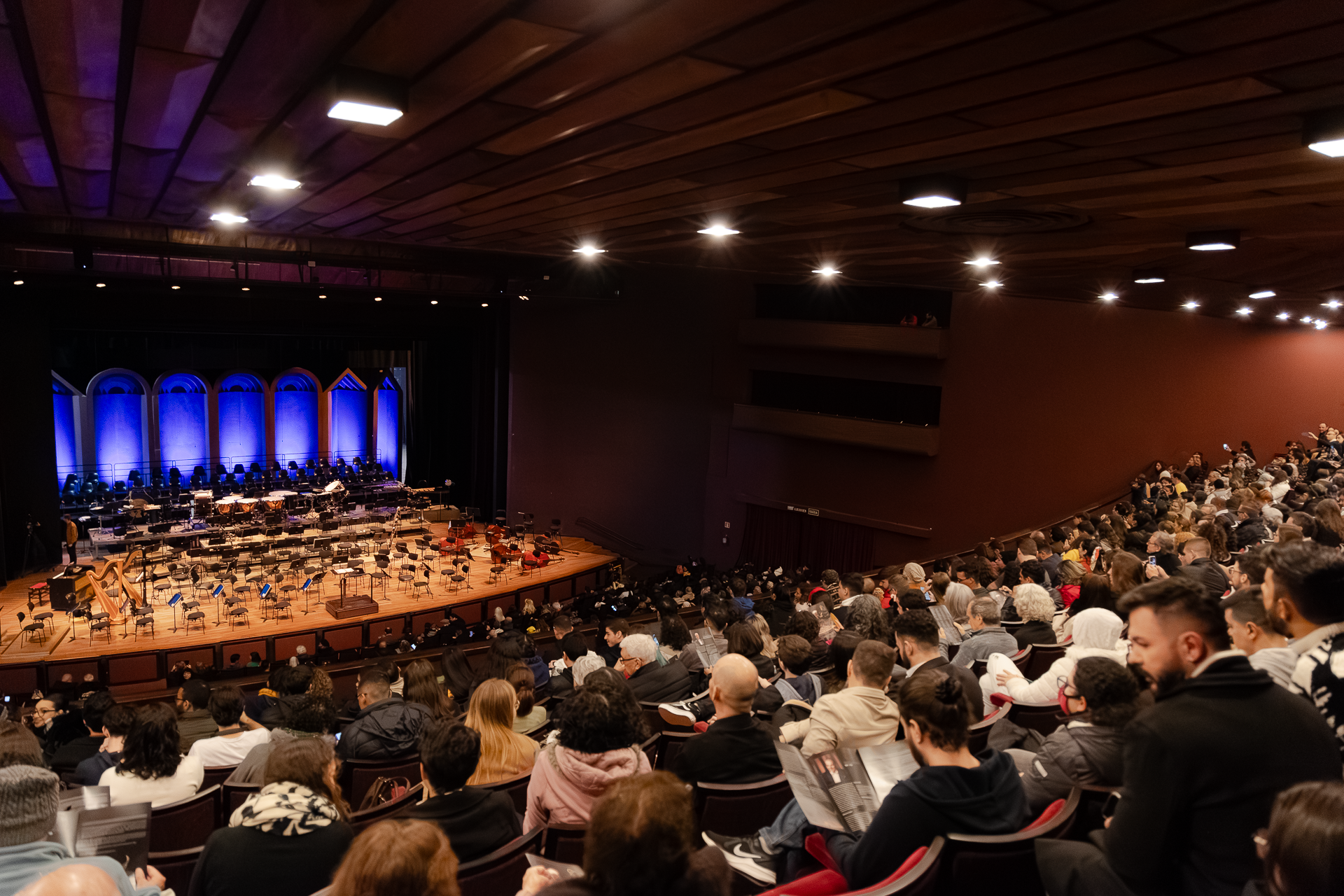 Pessoas sentadas na plateia do Guairão aguardam o início do concerto. No palco, todos os lugares dos músicos estão organizados e alguns instrumentos dispostos.