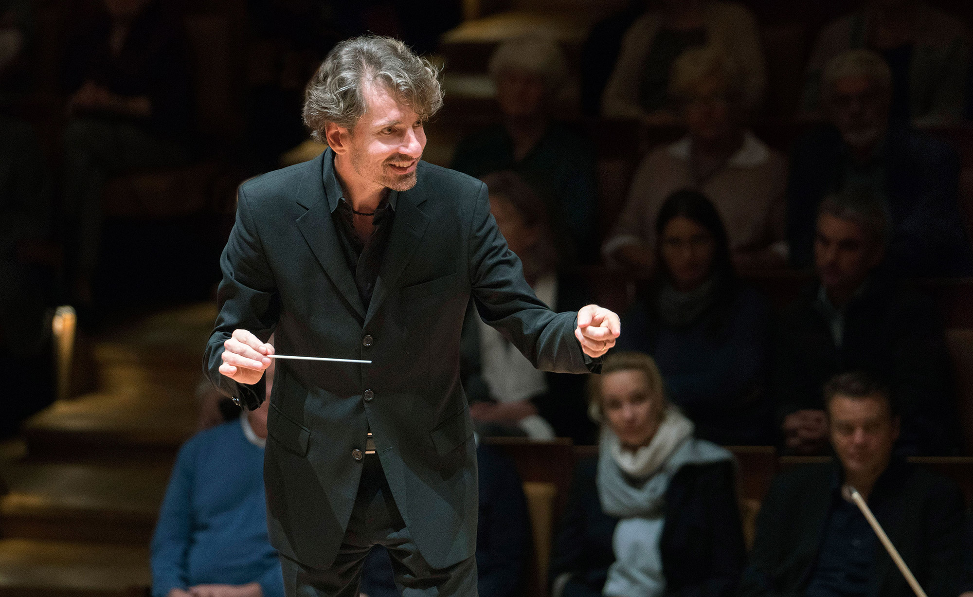 maestro regendo orquestra com sorriso no rosto. público assiste ao fundo