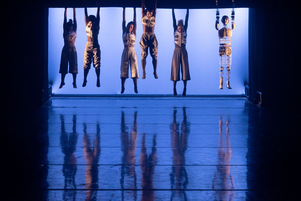 bailarinos pendem em cenário iluminado com luz azul. Do lado direito, boneco imita os bailarinos