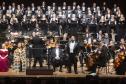 Toda a orquestra, com seus instrumentos, o coro, os solistas, os músicos convidados e o maestro estão posicionados durante o concerto no placo do Teatro Guaíra, com roupas de gala. 