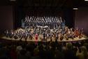 Toda a orquestra, com seus instrumentos, o coro, os solistas, os músicos convidados e o maestro estão posicionados durante o concerto no placo do Teatro Guaíra, com roupas de gala. 