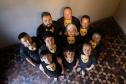Grupo teatral com máscaras abaixo do queixo e olhando para cima. A perspectiva mostra o chão de tacos com desenho de estrelas e uma parte da parede branca