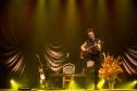 Nando Reis tocando violão no palco. Ao fundo, o cenário conta com um mesa, cadeira, vaso com plantas e outro violão em apoio