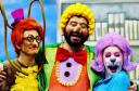 Personagens Doroteia, Nico e Ritinha sorrindo em frente a uma prateleira da biblioteca