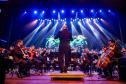 maestro à frente de Orquestra, luzes coloridas ao fundo ilustram o cenário do concerto