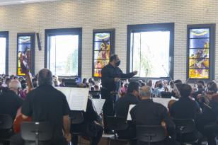 Foto dos músicos tocando dentro de uma igreja. Eles vestem preto. O maestro está em pé, regendo a orquestra.