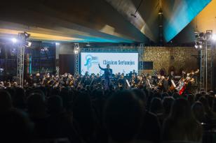 Foto do maestro conversando com o público. A orquestra está esperando para tocar ao fundo. Há um telão com o texto "Clássicos Sertanejos"