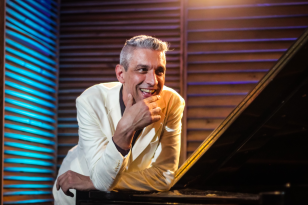 Foto de Thiago Lima apoiando seus braços em um piano. Ele veste uma camisa preta e terno branco. Seus cabelos são brancos e está com uma feição sorridente