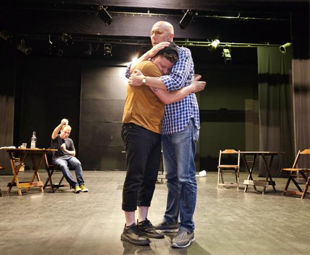 personagens abraçados em cena com mesas e cadeiras atrás