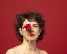 Adriana Calcanhoto aparece com uma rosa vermelha ao lado e, depois, na frente do rosto. No fundo, a cor vermelha também.