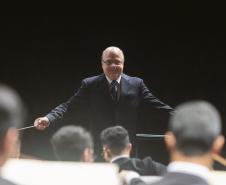 Toda a orquestra, com seus instrumentos, o coro, os solistas, os músicos convidados e o maestro estão posicionados durante o concerto no placo do Teatro Guaíra, com roupas de gala. 