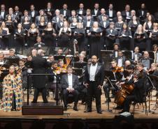 Toda a orquestra, com seus instrumentos, o coro, os solistas, os músicos convidados e o maestro estão posicionados durante o concerto no placo do Teatro Guaíra, com roupas de gala. 