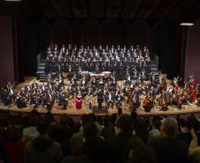 Toda a orquestra, com seus instrumentos, o coro, os solistas, os músicos convidados e o maestro estão posicionados durante o concerto no placo do Teatro Guaíra, com roupas de gala. 