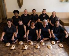 grupo reunido em uma sala com parede meia branca e meia amarela, usando roupas pretas e mostrando as máscaras no chão de tacos.