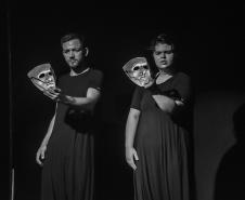 vestidos de preto, dois atores seguram máscara com a mão esquerda. foto em preto e branco