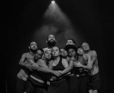 foto em preto e branco. 10 personagens abraçados sob fundo preto com iluminação direta acima dos atores