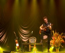 Nando Reis tocando violão no palco. Ao fundo, o cenário conta com um mesa, cadeira, vaso com plantas e outro violão em apoio