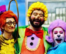 Personagens Doroteia, Nico e Ritinha sorrindo em frente a uma prateleira da biblioteca