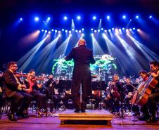 maestro à frente de Orquestra, luzes coloridas ao fundo ilustram o cenário do concerto