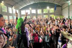 A plateia formada por crianças interage com a banda. Os integrantes usam roupas e acessórios coloridos, que destacam o universo lúdico das crianças.