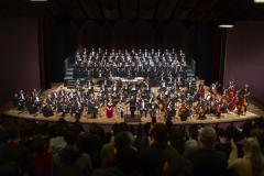 Toda a orquestra, com seus instrumentos, o coro, os solistas, os músicos convidados e o maestro estão posicionados durante o concerto no placo do Teatro Guaíra, com roupas de gala. 