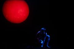 Cena da peça, com lua vermelha e mulher em luz azul.