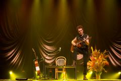 Nando Reis tocando violão no palco. Ao fundo, o cenário conta com um mesa, cadeira, vaso com plantas e outro violão em apoio