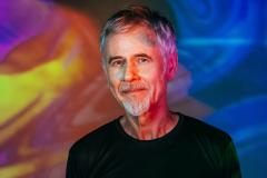 Ritchie sorri, de camisa preta em frente a um fundo colorido