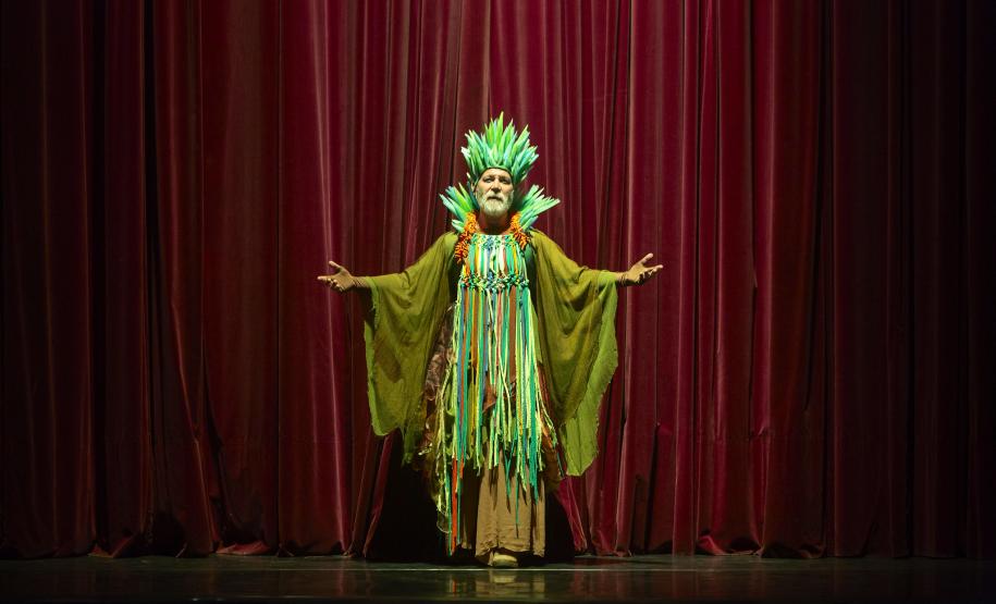 Ator Ranieri Gonzalez com a roupa verde do Senhor das Lendas de pé no paco do Guaíra. Ao fundo a cortina vermelha.