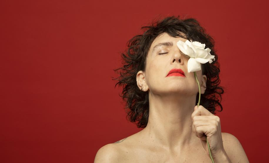 Adriana Calcanhoto aparece com uma rosa vermelha ao lado e, depois, na frente do rosto. No fundo, a cor vermelha também.