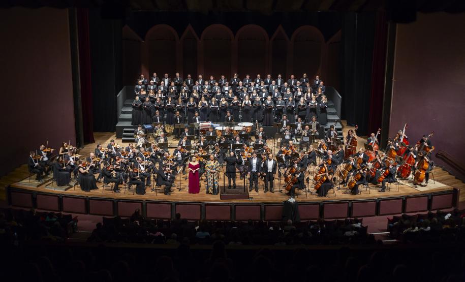 Toda a orquestra, com seus instrumentos, o coro, os solistas, os músicos convidados e o maestro estão posicionados durante o concerto no placo do Teatro Guaíra, com roupas de gala.