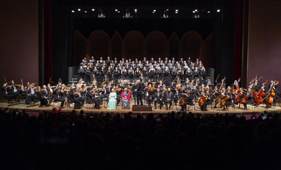 Toda a orquestra, com seus instrumentos, o coro, os solistas, os músicos convidados e o maestro estão posicionados durante o concerto no placo do Teatro Guaíra, com roupas de gala.