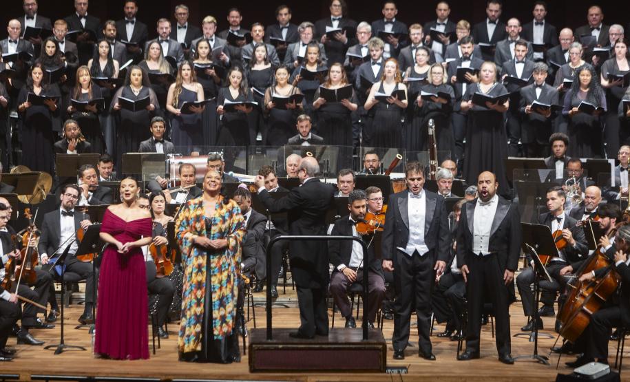 Toda a orquestra, com seus instrumentos, o coro, os solistas, os músicos convidados e o maestro estão posicionados durante o concerto no placo do Teatro Guaíra, com roupas de gala.