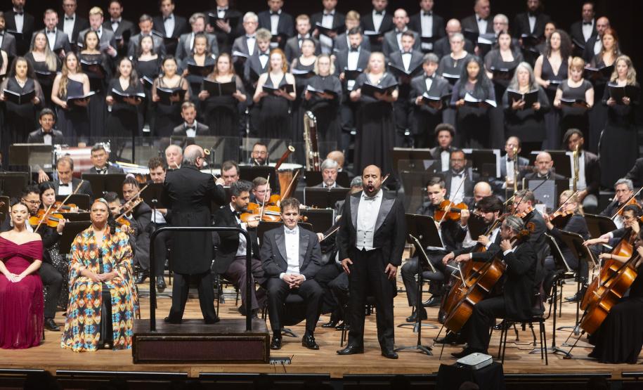 Toda a orquestra, com seus instrumentos, o coro, os solistas, os músicos convidados e o maestro estão posicionados durante o concerto no placo do Teatro Guaíra, com roupas de gala.