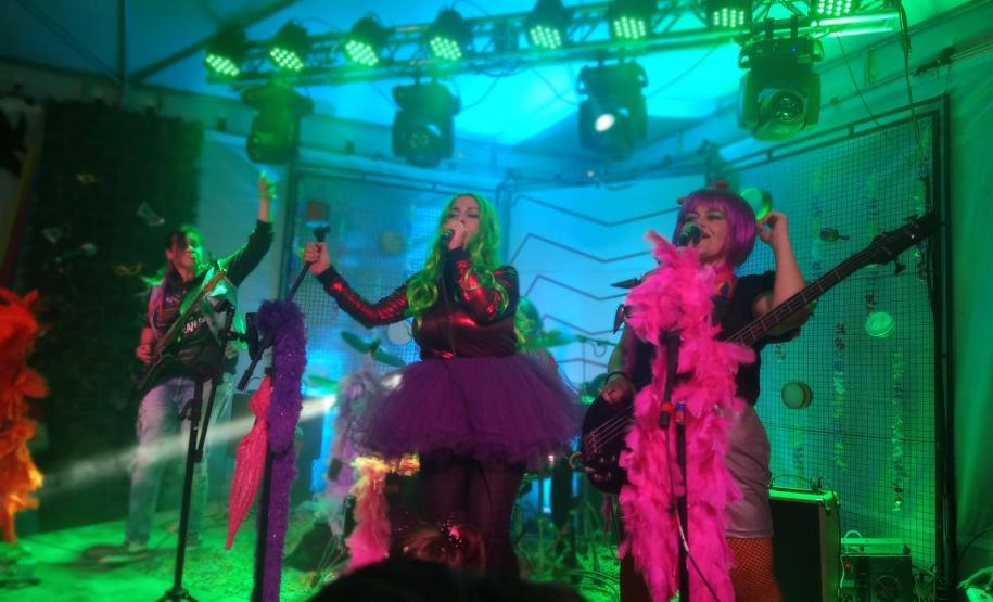 A plateia formada por crianças interage com a banda. Os integrantes usam roupas e acessórios coloridos, que destacam o universo lúdico das crianças.