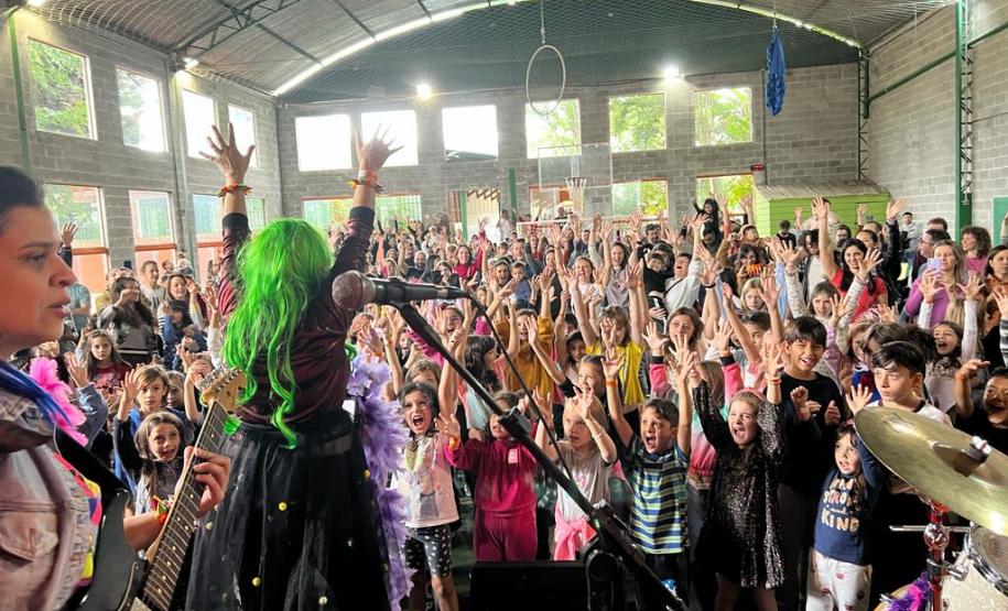 A plateia formada por crianças interage com a banda. Os integrantes usam roupas e acessórios coloridos, que destacam o universo lúdico das crianças.