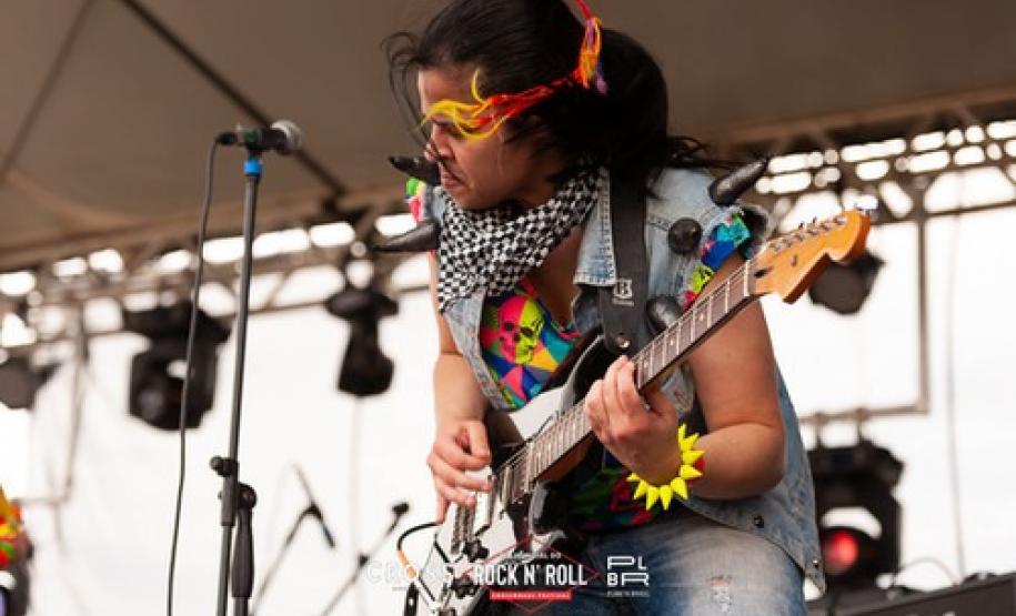 A plateia formada por crianças interage com a banda. Os integrantes usam roupas e acessórios coloridos, que destacam o universo lúdico das crianças.