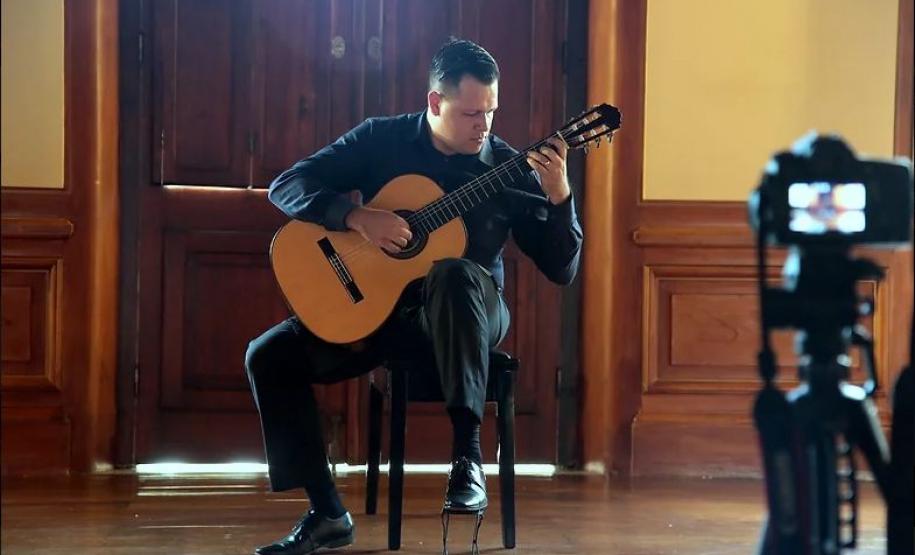 Walmor Boza tocando violão numa sala enquanto uma câmera está à sua frente