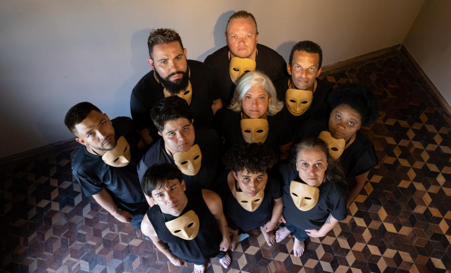 Grupo teatral com máscaras abaixo do queixo e olhando para cima. A perspectiva mostra o chão de tacos com desenho de estrelas e uma parte da parede branca